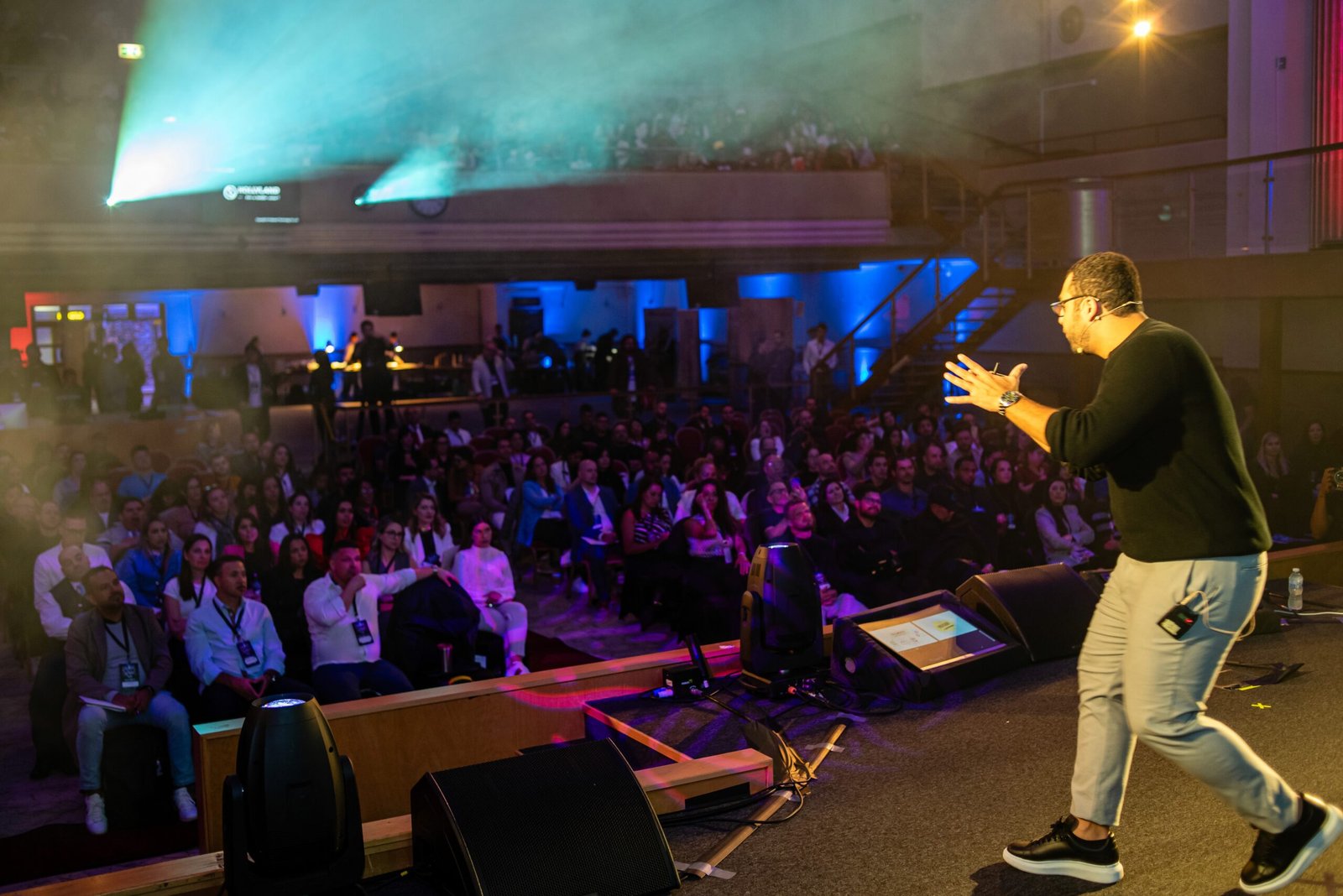 A speaker addressing a seated audience from a stage lit in blue