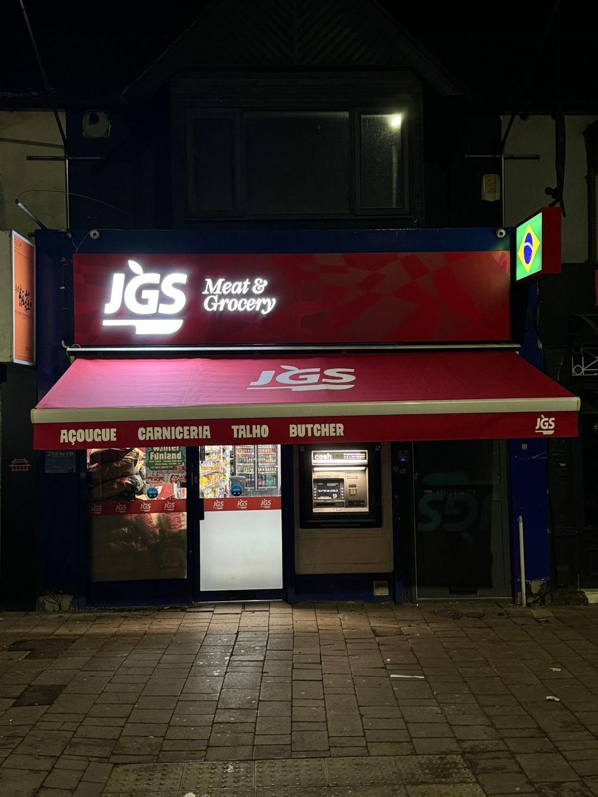 A night photograph of the JGS storefront, its red awning listing the counters inside