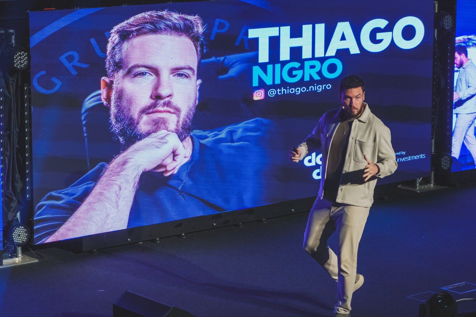A speaker on stage in front of a large LED screen showing Thiago Nigro